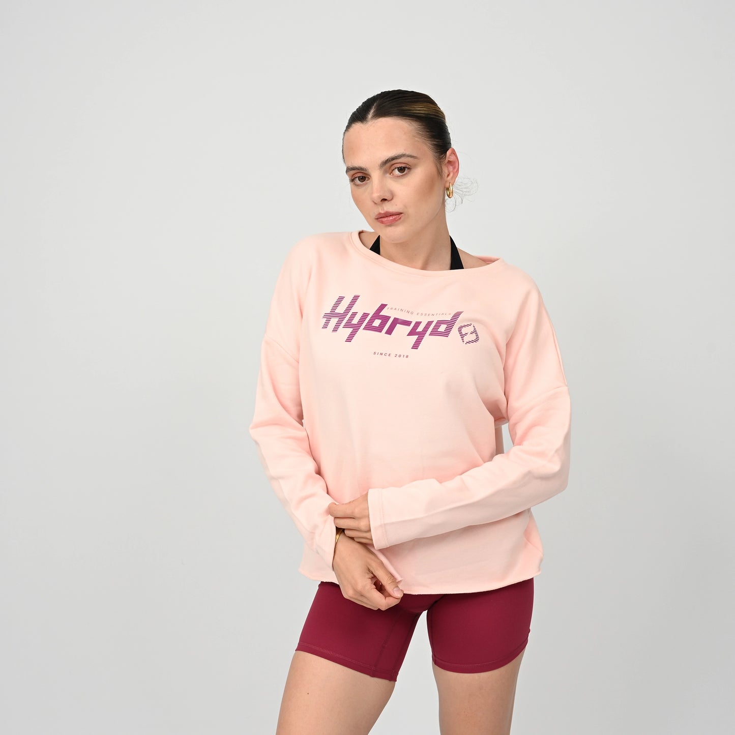 Woman wearing a pink sweatshirt with 'Hybryd' logo and maroon shorts on a grey background