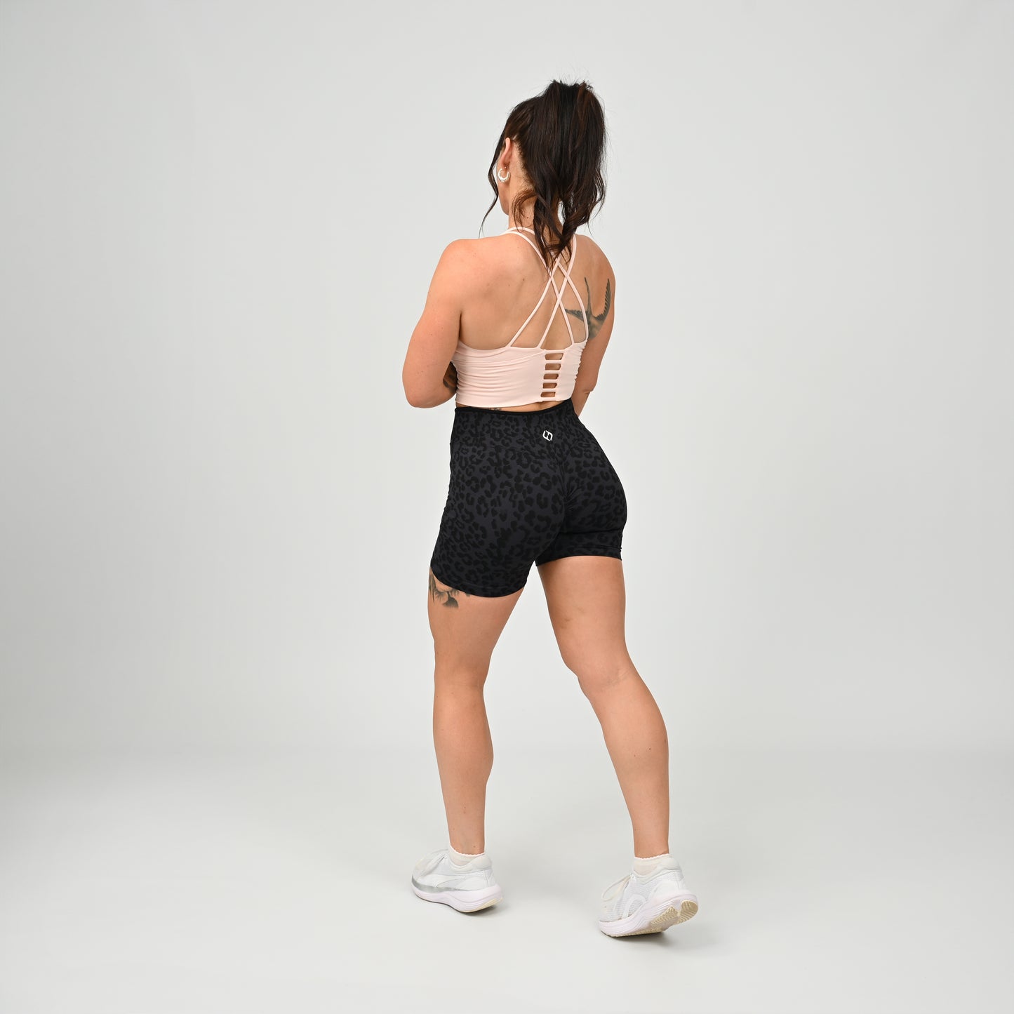 A woman is shown from behind wearing a light pink Hybryd athletic sports bra with crisscross straps and black patterned high-waisted gym shorts, paired with white sneakers, against a light gray studio background.