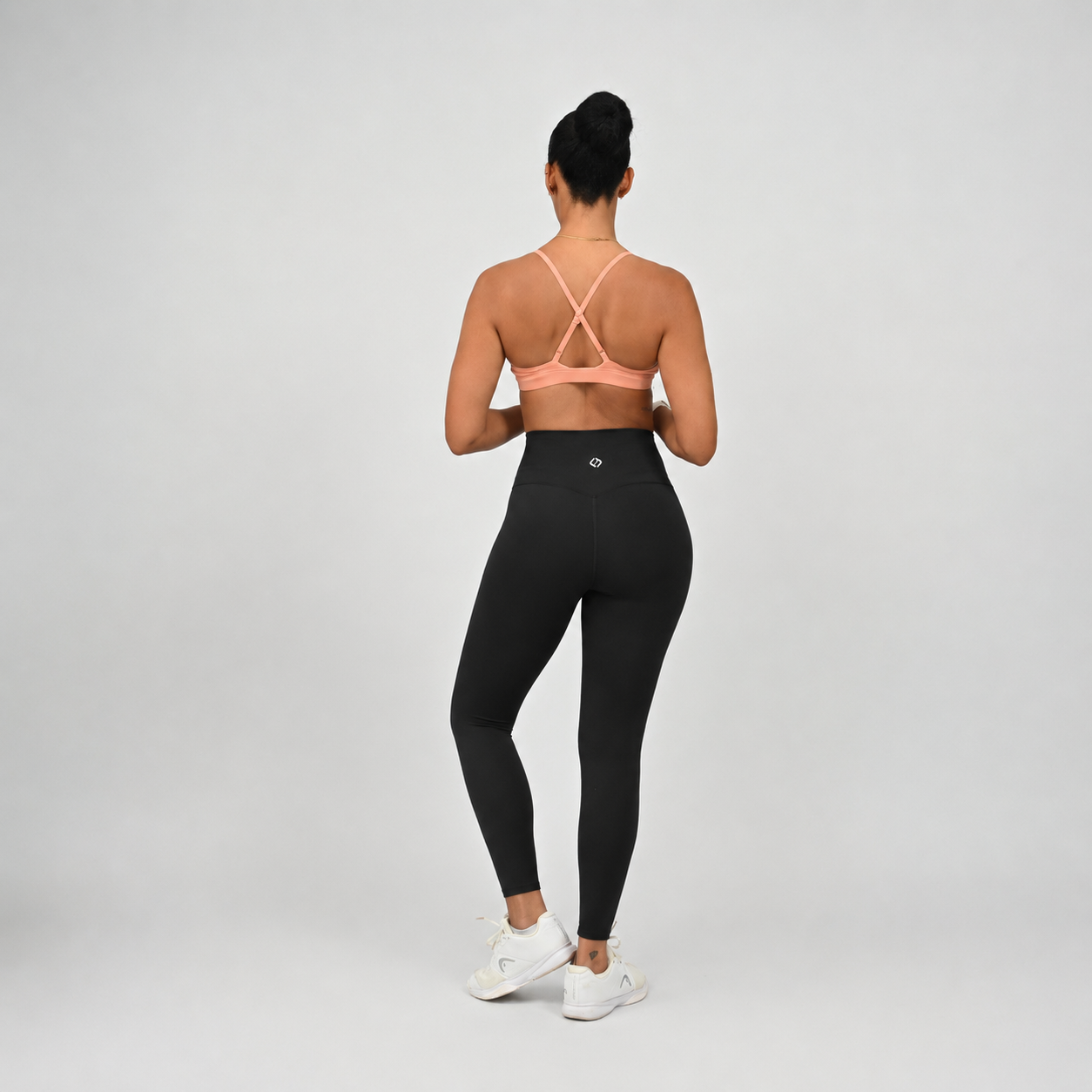 A woman is shown from behind wearing black high-waisted leggings and a peach-pink sports bra with adjustable straps, standing against a light gray studio background.