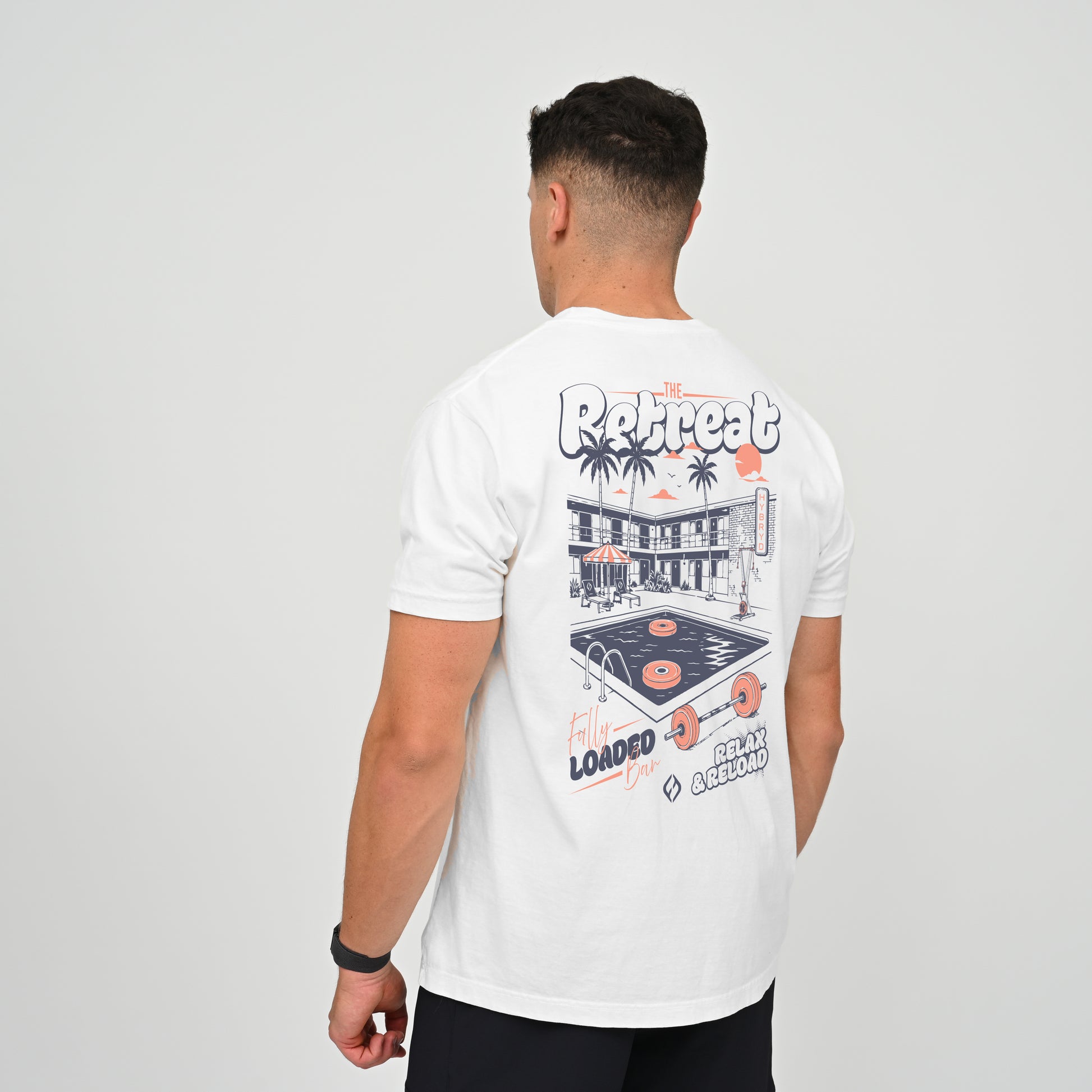 A crossfit athlete wearing a white graphic t-shirt, viewed from behind to show the back print. The graphic features a retro diner, palm trees, a vinyl record, and text reading Hybryd Retreat.