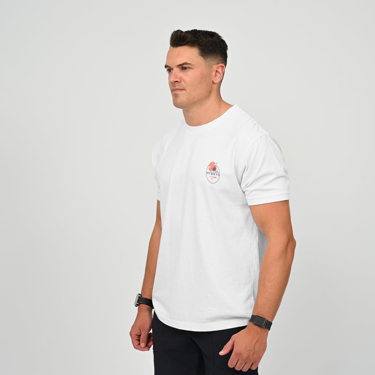 A man wearing a white short-sleeve crewneck t-shirt with a small circular graphic on the left chest, paired with dark pants, standing against a light gray studio background. Both items from the gymwear brand Hybryd.
