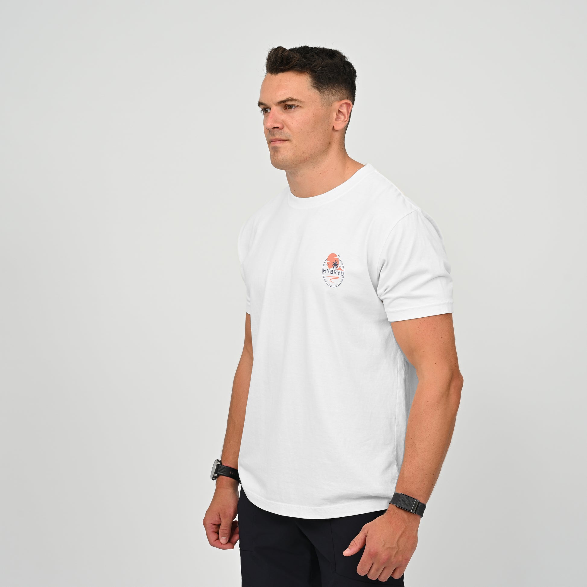 A man wearing a white short-sleeve crewneck t-shirt with a small circular graphic on the left chest, paired with dark pants, standing against a light gray studio background. Both items from the gymwear brand Hybryd.