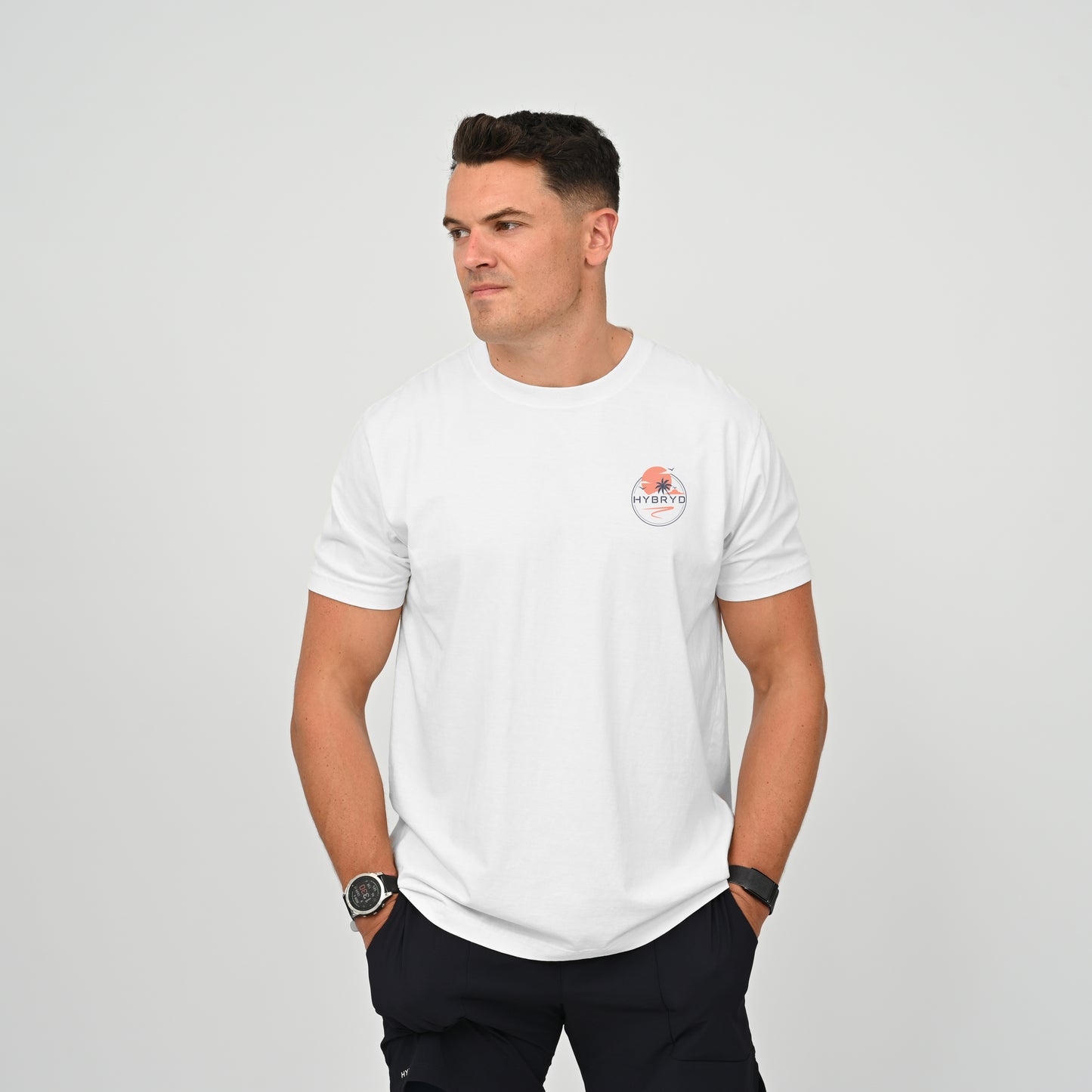 Hyrox athlete wearing a white crew-neck t-shirt with a small circular Hybryd logo on the left chest, paired with dark pants, standing against a light gray studio background. From activewear brand Hybryd.