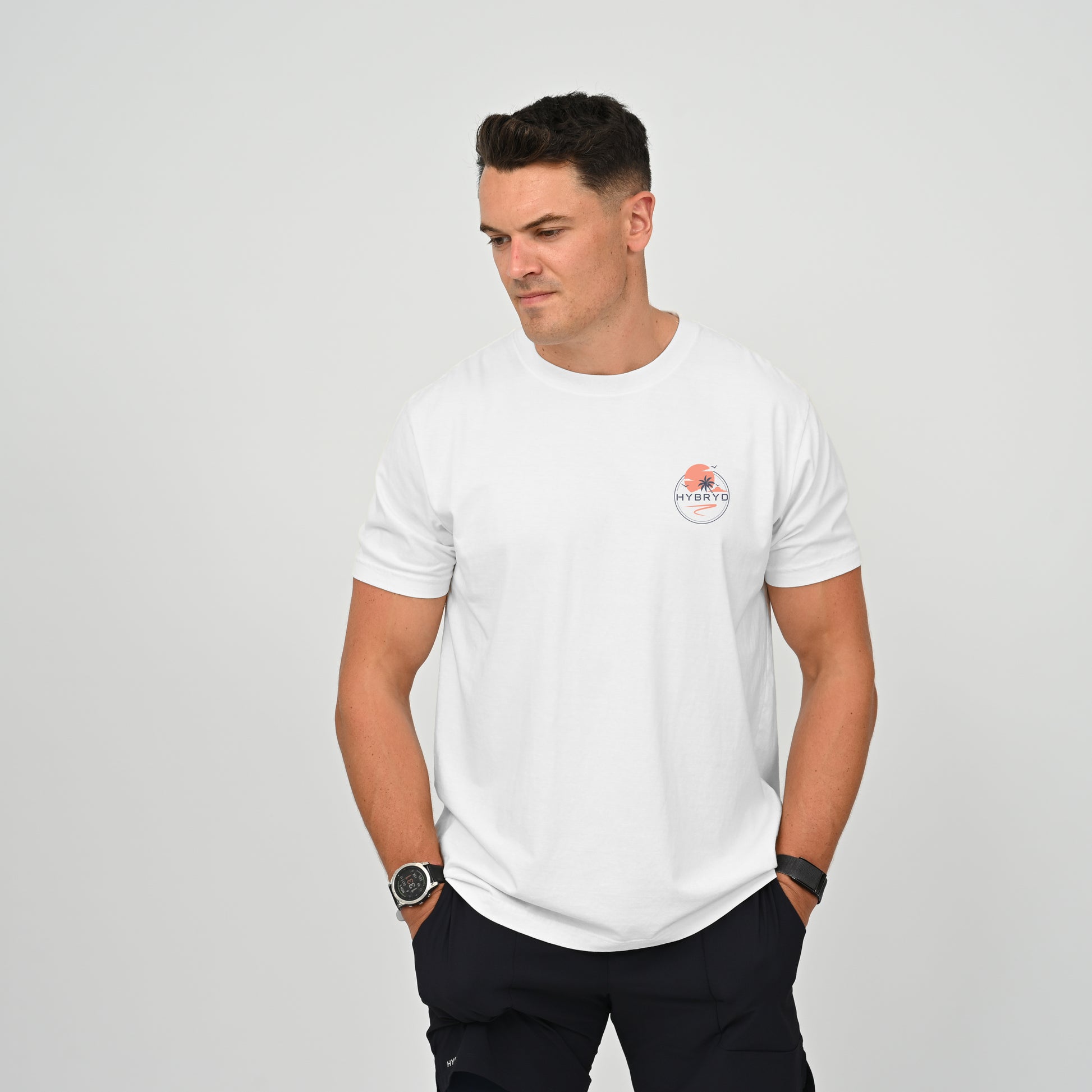 A man wearing a white crew-neck t-shirt with a small circular Hybryd logo on the left chest, paired with dark pants, standing against a light gray studio background. From activewear brand Hybryd.