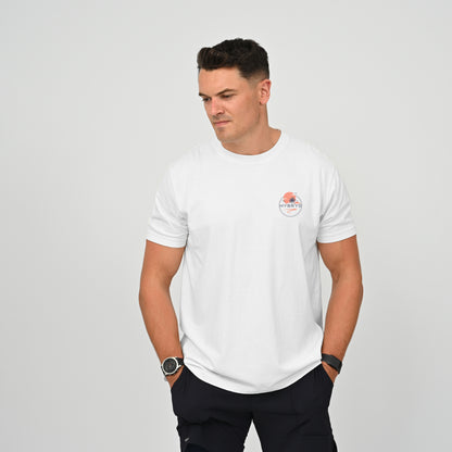 A man wearing a white crew-neck t-shirt with a small circular Hybryd logo on the left chest, paired with dark pants, standing against a light gray studio background. From activewear brand Hybryd.