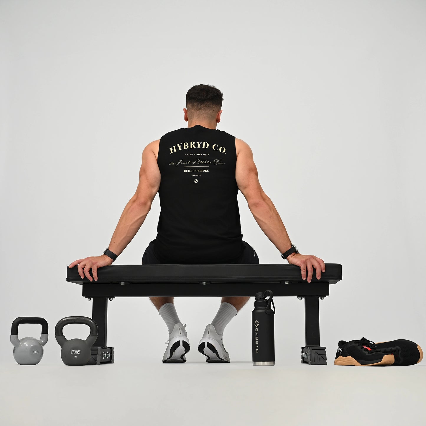 A man sits on a black weight bench viewed from behind, wearing a black HYBRYD CO sleeveless gym vest and black shorts. The scene is set in a minimalist studio with a gray background, featuring a kettlebell on the left, a water bottle, and a black and tan athletic shoe on the right.