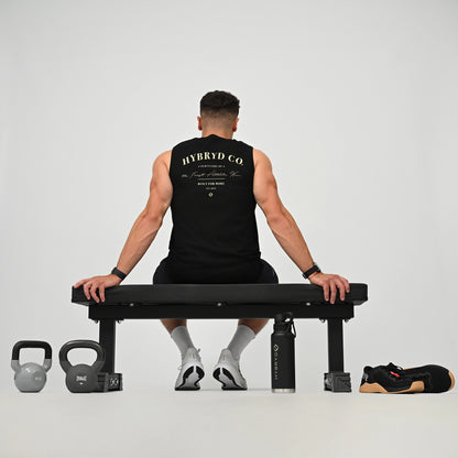 A man sits on a black weight bench viewed from behind, wearing a black HYBRYD CO sleeveless gym vest and black shorts. The scene is set in a minimalist studio with a gray background, featuring a kettlebell on the left, a water bottle, and a black and tan athletic shoe on the right.