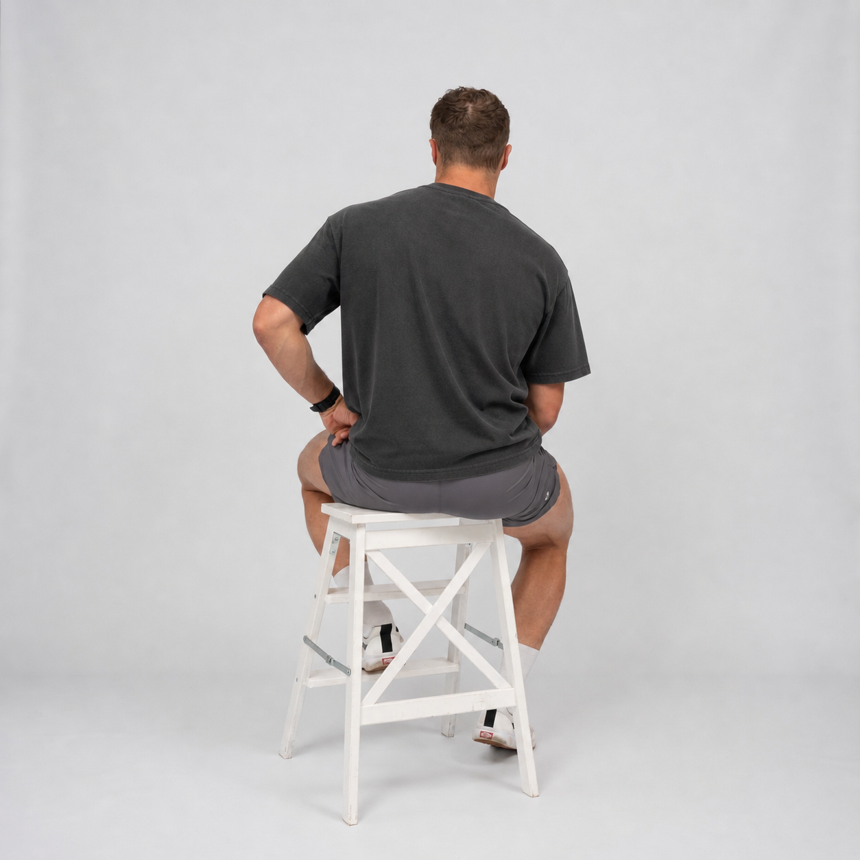 Man sitting on a white stool wearing a Hybryd t-shirt and  shorts against a light grey background