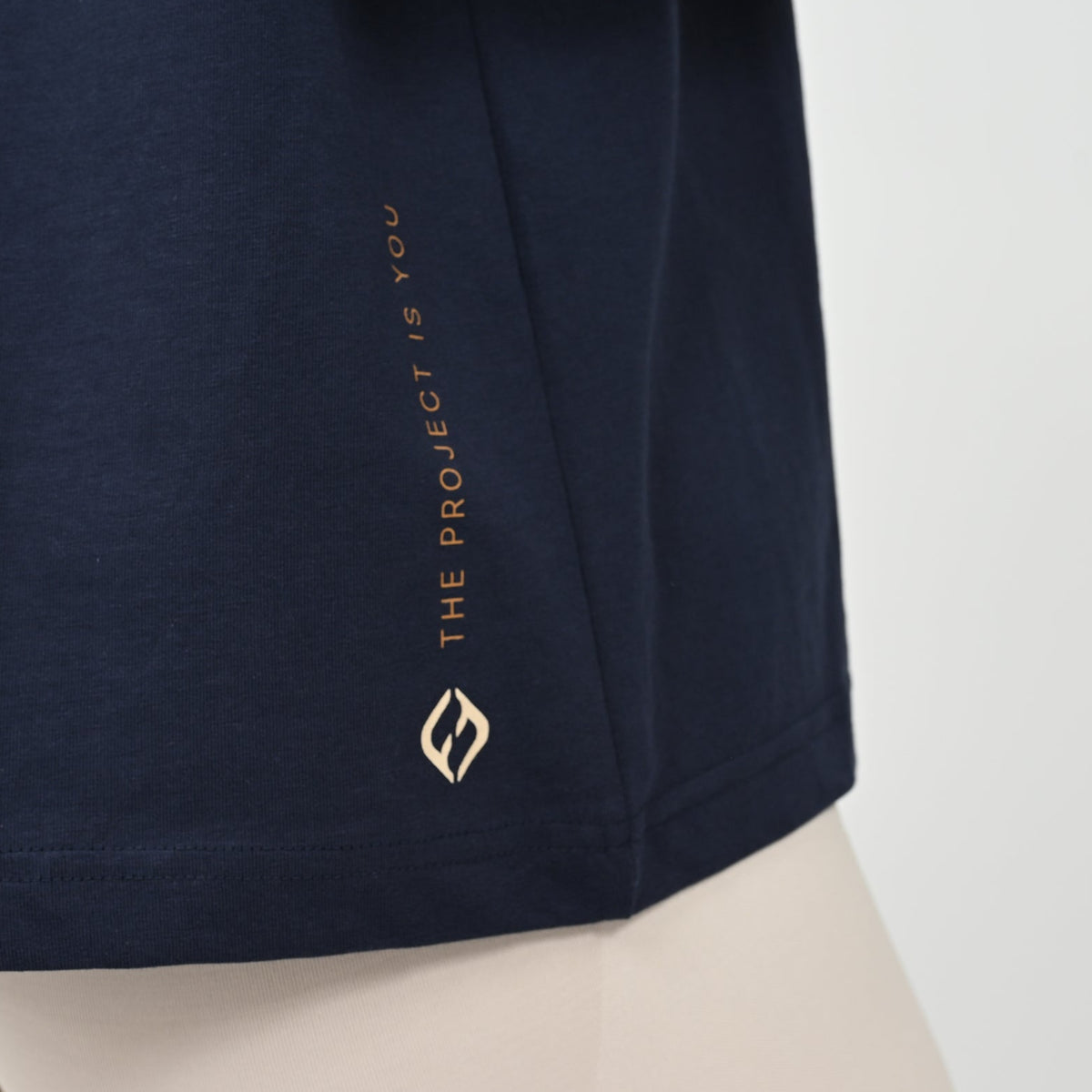 Close-up of a navy blue garment, featuring vertical gold text reading 'THE PROJECT IS YOU' and a matching geometric Hybryd logo near the hem, displayed on a gum person against a light background.