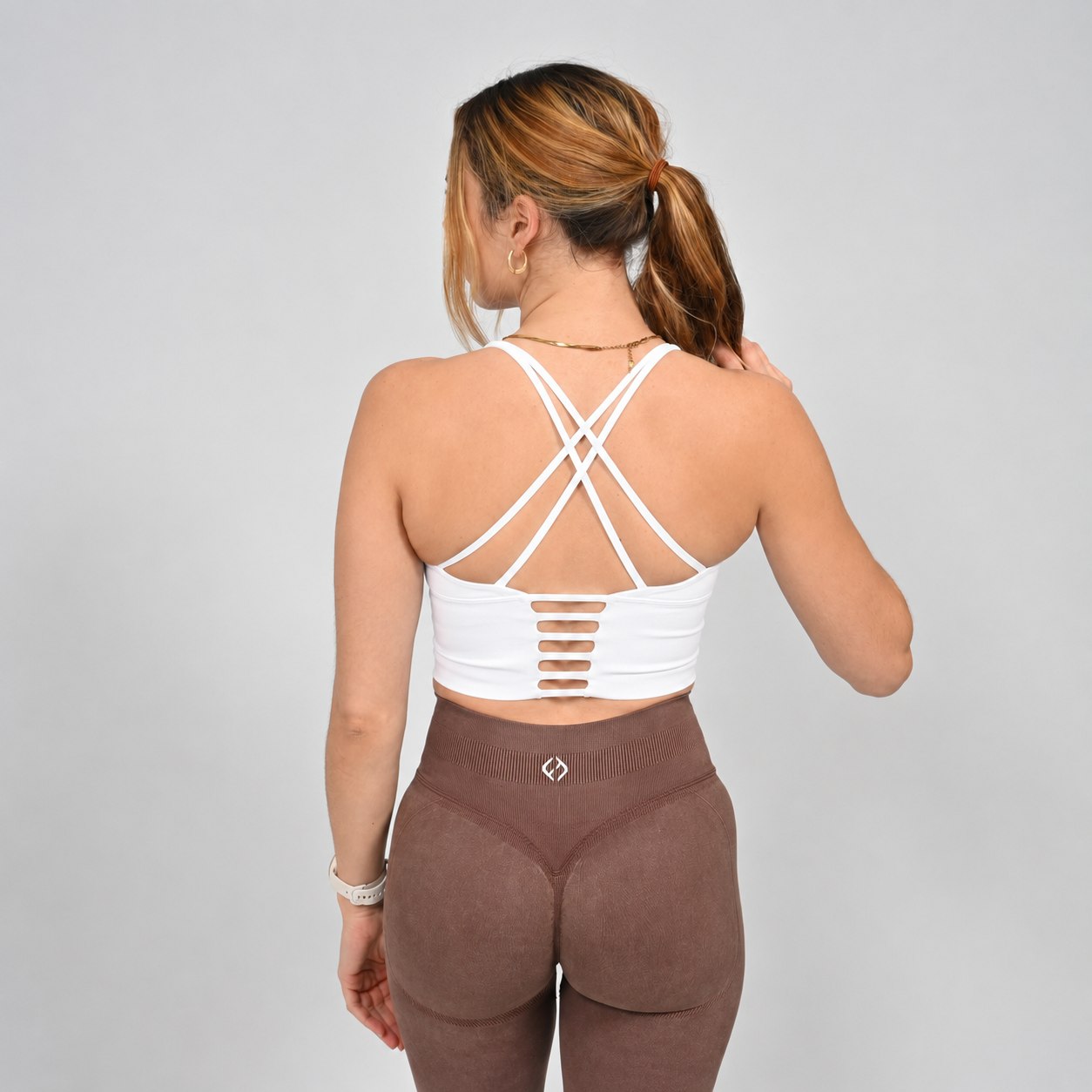 A Hyrox athlete wearing a white long line sports bra with brown leggings.  Pictured from the rear showing off the strappy back. Made by the gymwear brand Hybryd.