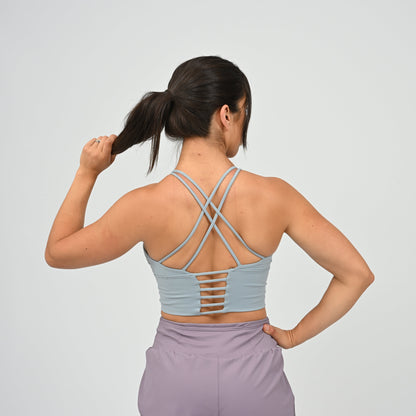 A woman modeling a light blue Hybryd sports bra with a small white logo on the chest, paired with lavender run shorts, against a light gray studio background. Shot from behind
