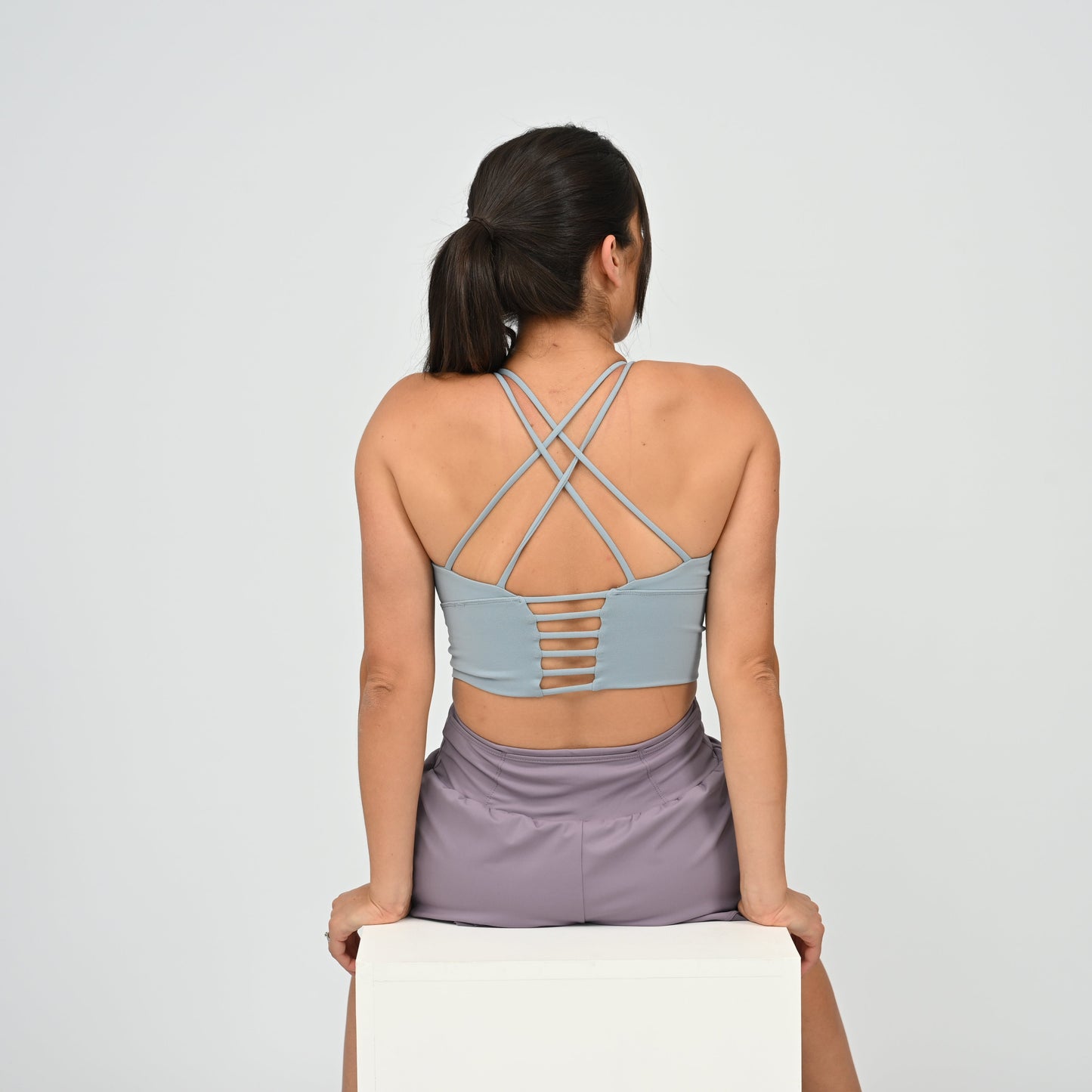A woman modeling a light blue Hybryd sports bra with a strappy back,  paired with lavender run shorts, against a light gray studio background.