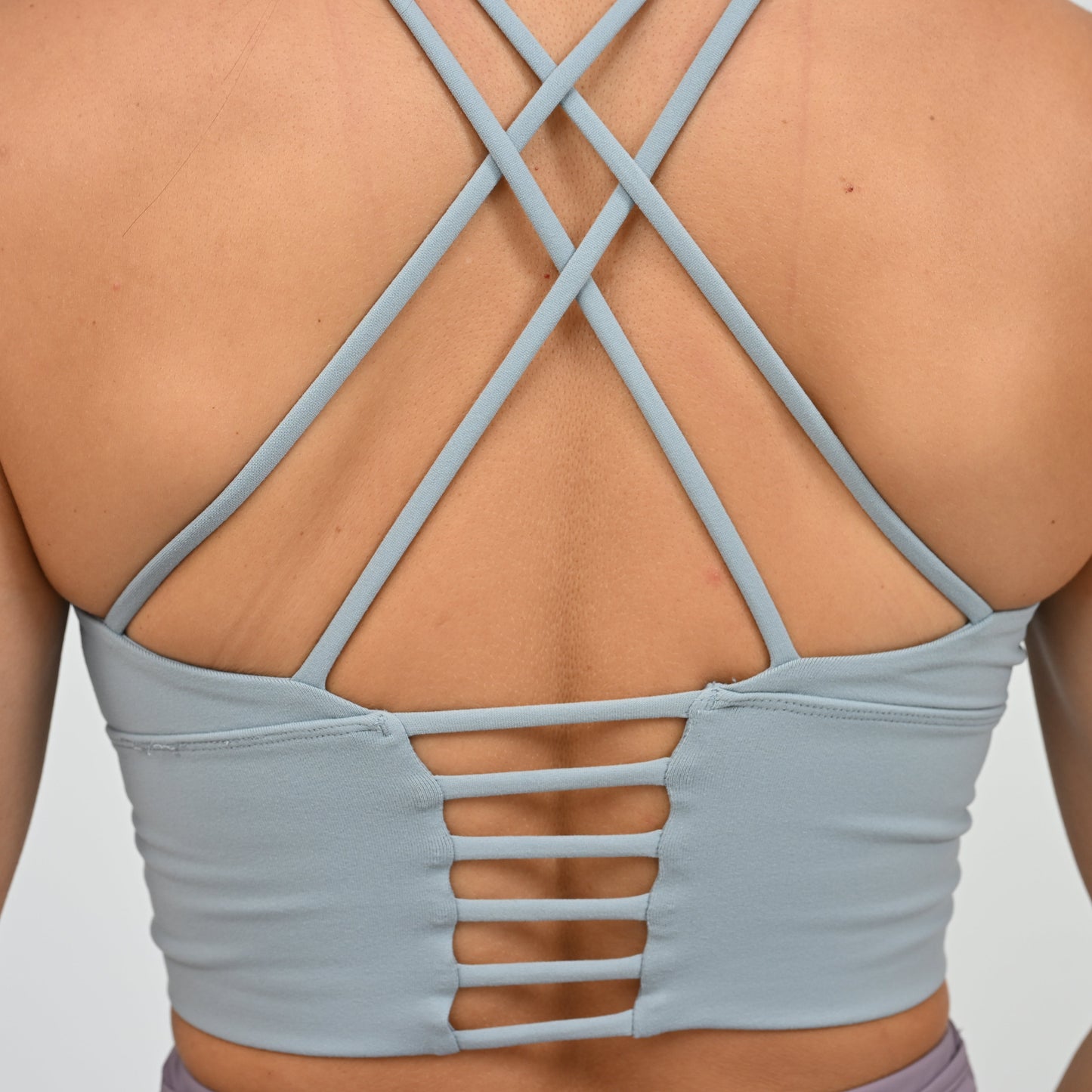 Close-up back view of a person wearing a light blue-gray strappy sports bra  from Hybryd with crisscross straps and cutout details at the lower back.