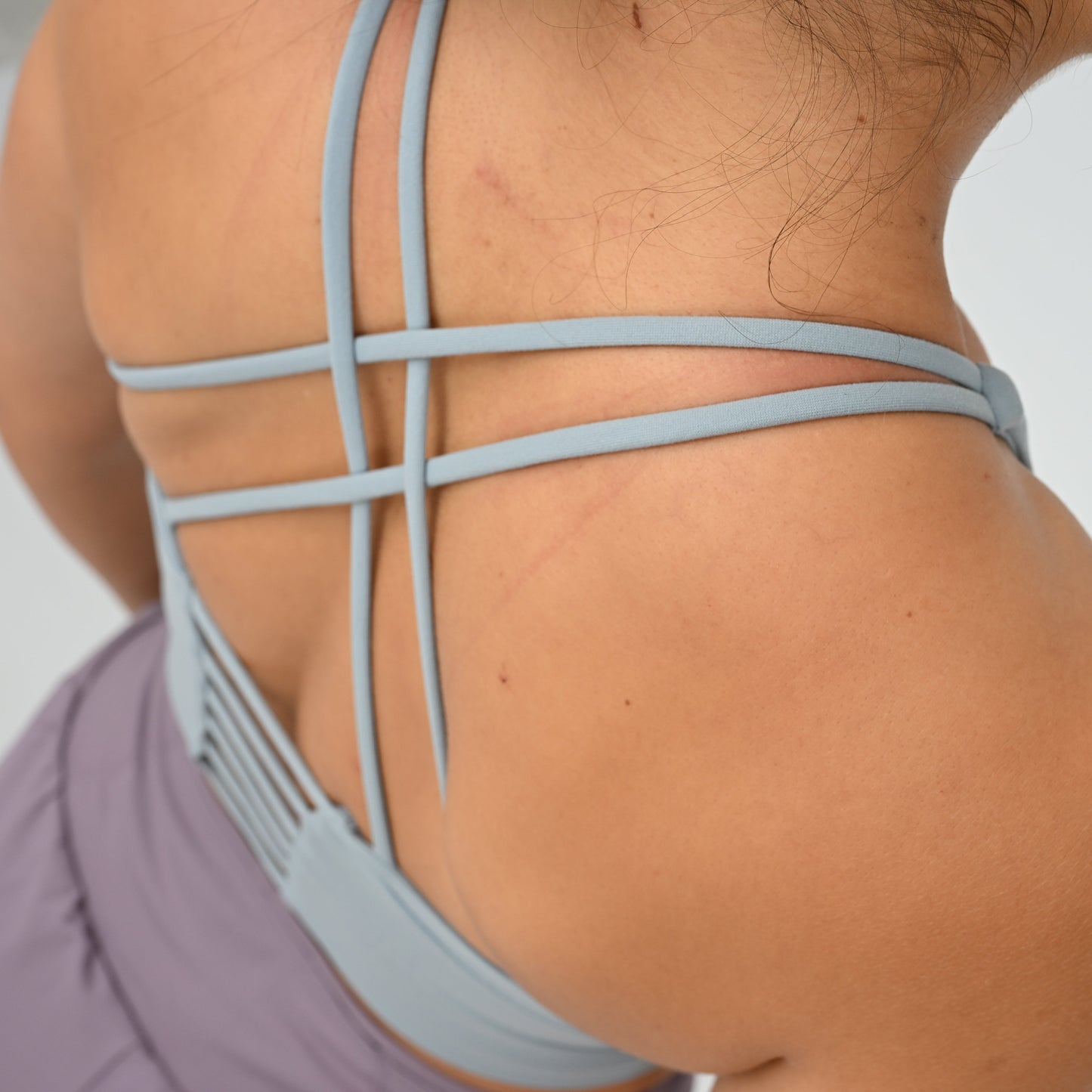 Close-up back view of a person wearing a light blue Hybryd strappy sports bra or tank top with a crisscross back design and a lavender sports bottom.