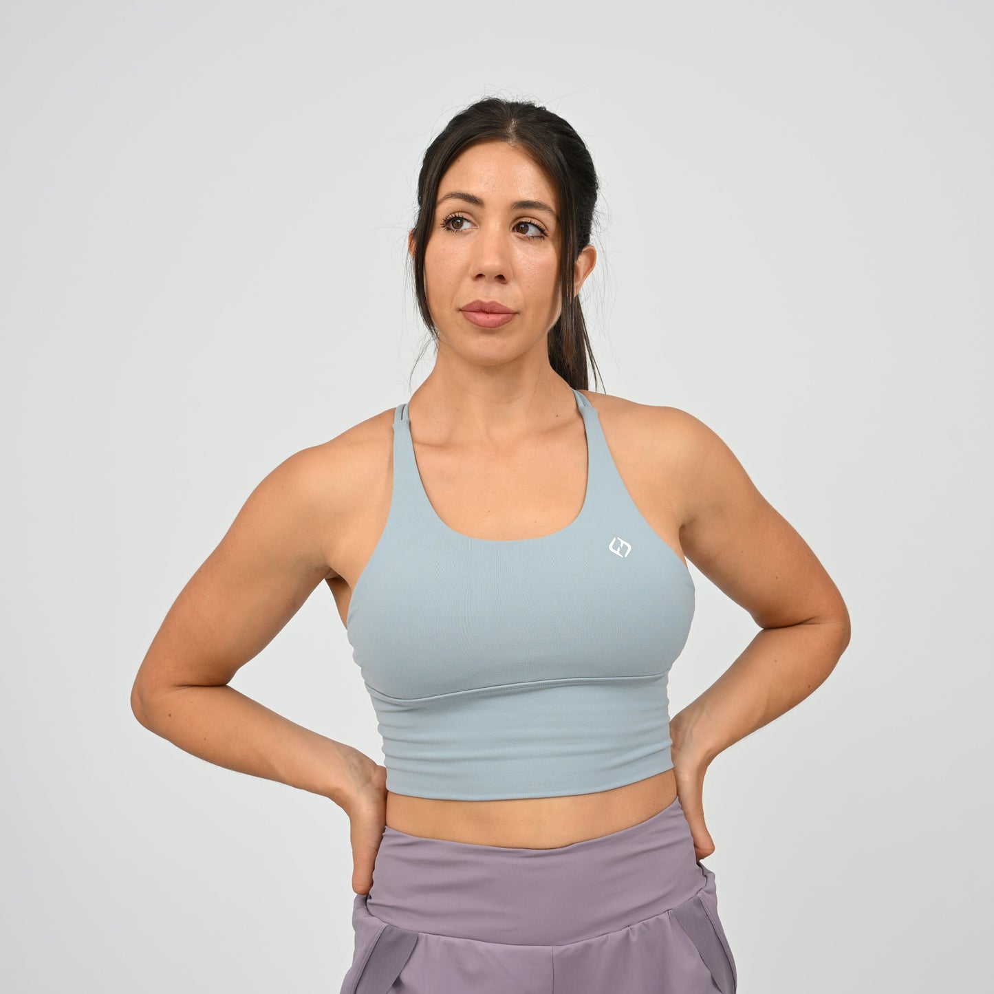 A woman modeling a light blue Hybryd sports bra with a small white logo on the chest, paired with lavender run shorts, against a light gray studio background.
