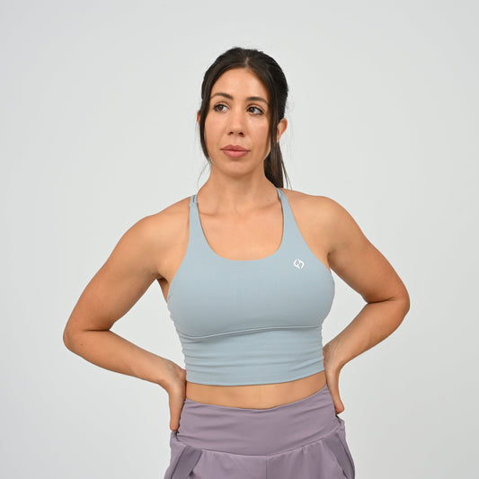 A woman modeling a light blue Hybryd sports bra with a small white logo on the chest, paired with lavender run shorts, against a light gray studio background.