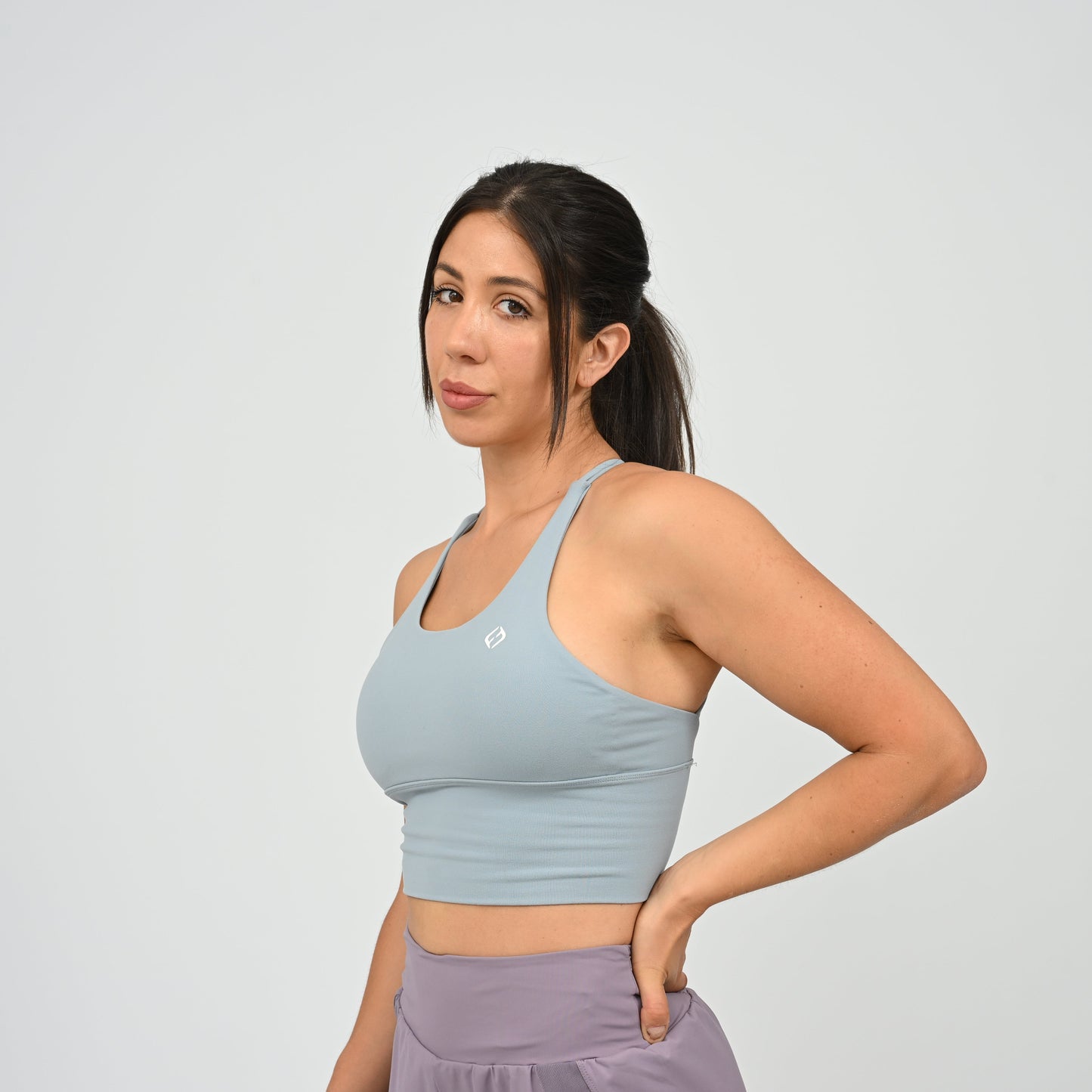 A woman modeling a light blue Hybryd sports bra with a small white logo on the chest, paired with lavender run shorts, against a light gray studio background.