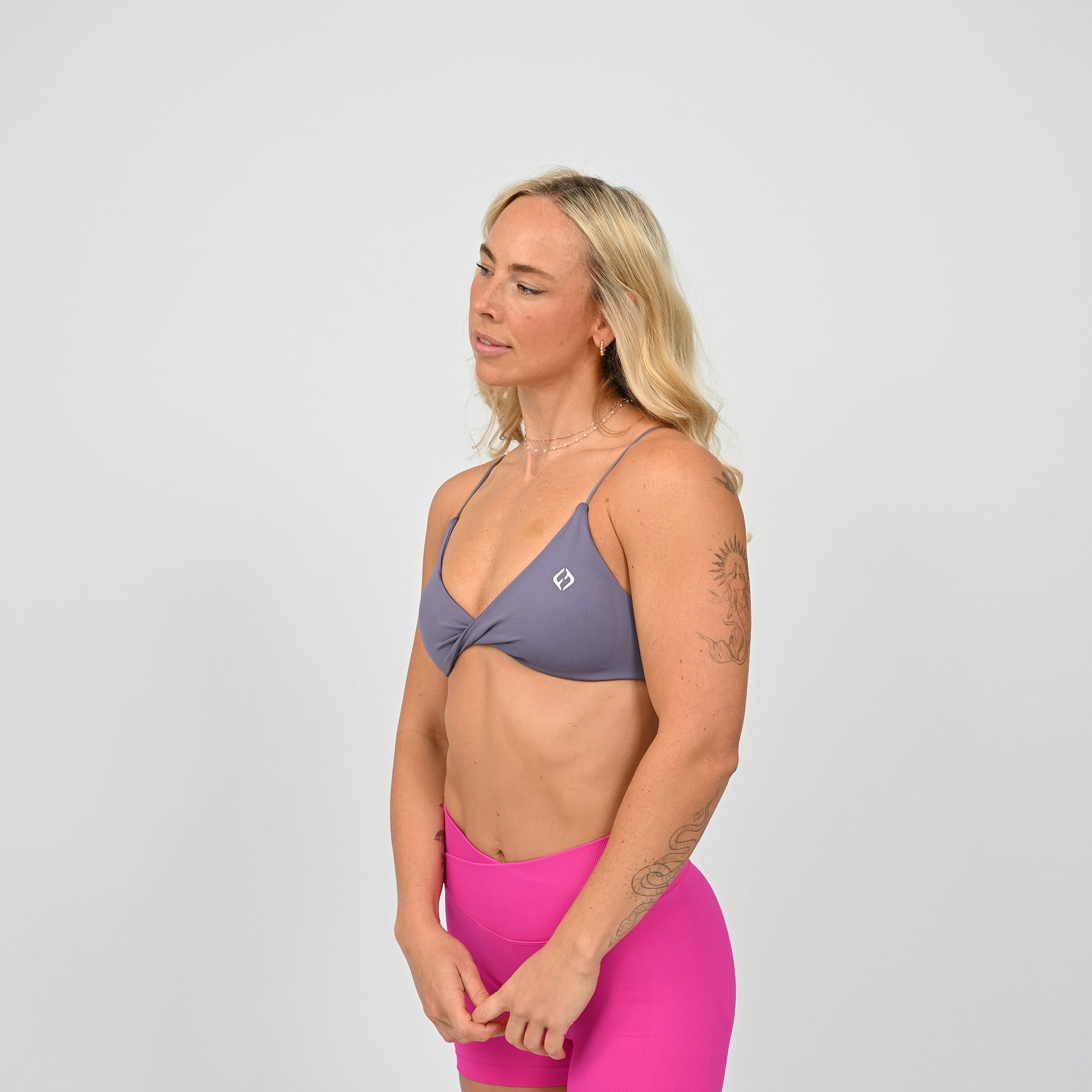 Gym girl wearing a grey minimal sports bra with crossover front. Worn with scrunch bum pink bootie shorts that have a crossoer waistband.  Made by the activewear brand Hybryd.