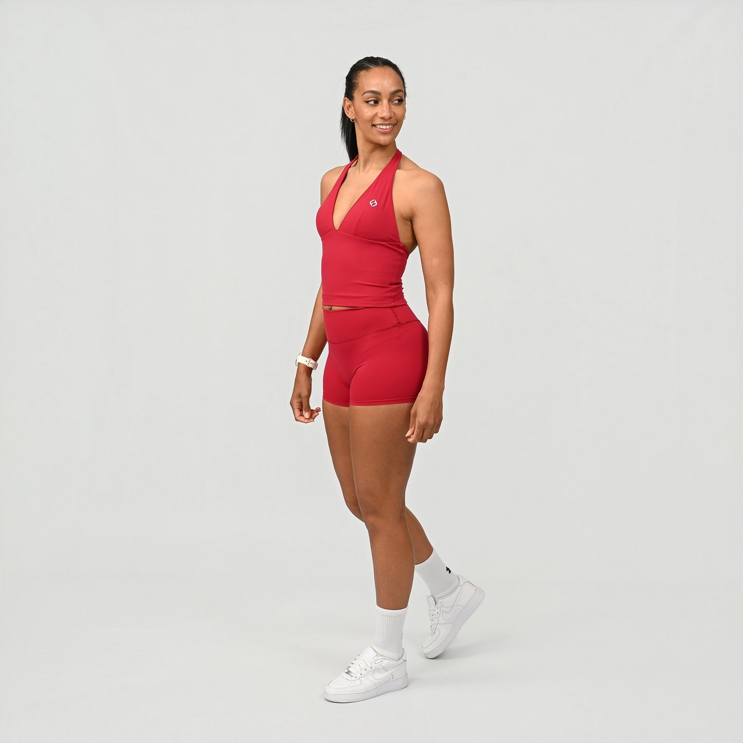 Woman wearing a Hybryd red athletic booty shorts and vest outfit on a light gray background. Worn by a HYROX athlete.