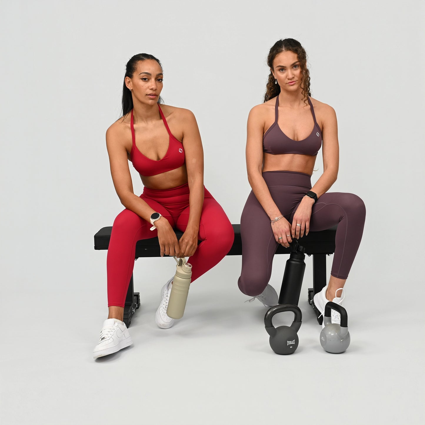 Two women in athletic wear matching gym sets from Hybryd, sitting on a bench with kettlebells. 