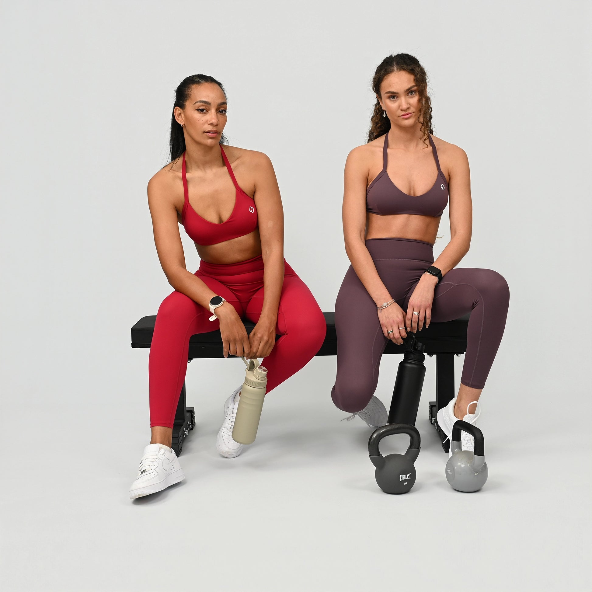Two women in athletic wear matching gym sets from Hybryd, sitting on a bench with kettlebells. 