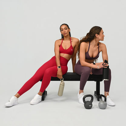 Two women in Hybryd athletic wear sitting on a bench with fitness equipment around them.