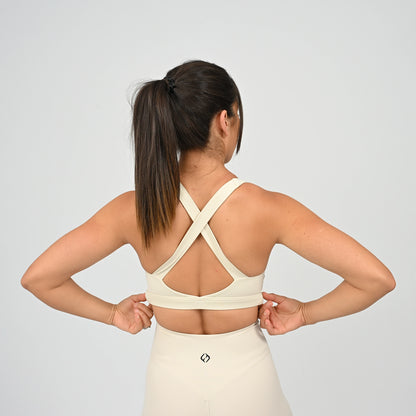 A woman shown from the back wearing a cream-colored sports bra with crisscross straps and matching high-waisted shorts, set against a plain light gray studio background. From activewear brand Hybryd.