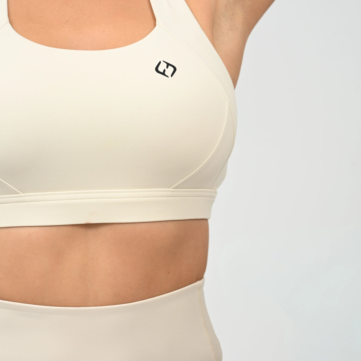 Close-up of a person wearing a cream-colored sports bra and matching high-waisted bottoms against a light gray studio background. A small black Hybryd logo is visible on the upper chest of the sports bra. From activewear brand Hybryd.