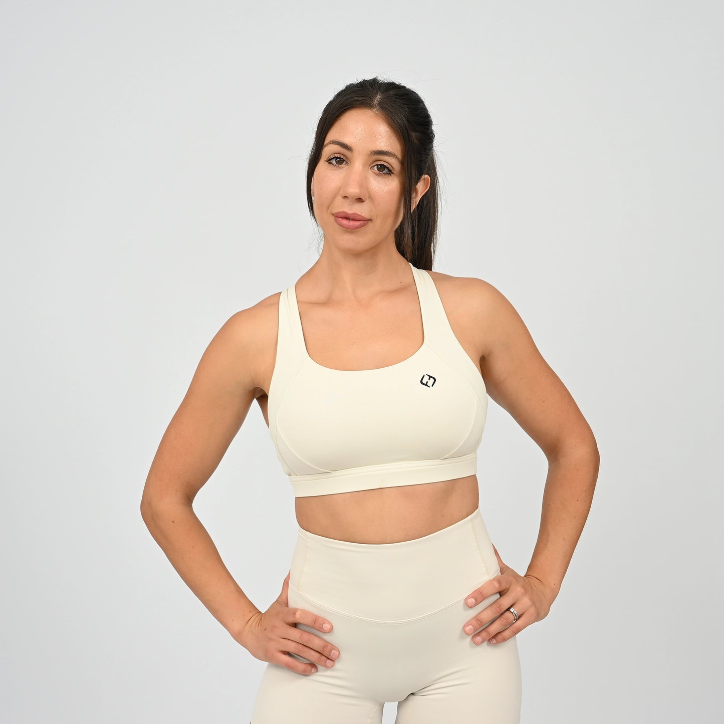 A woman modeling a matching cream-colored activewear set consisting of a scoop-neck sports bra and high-waisted shorts, set against a plain light gray studio background. From gymwear brand Hybryd.