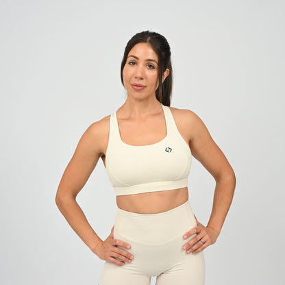 A woman modeling a matching cream-colored activewear set consisting of a scoop-neck sports bra and high-waisted shorts, set against a plain light gray studio background. From gymwear brand Hybryd.
