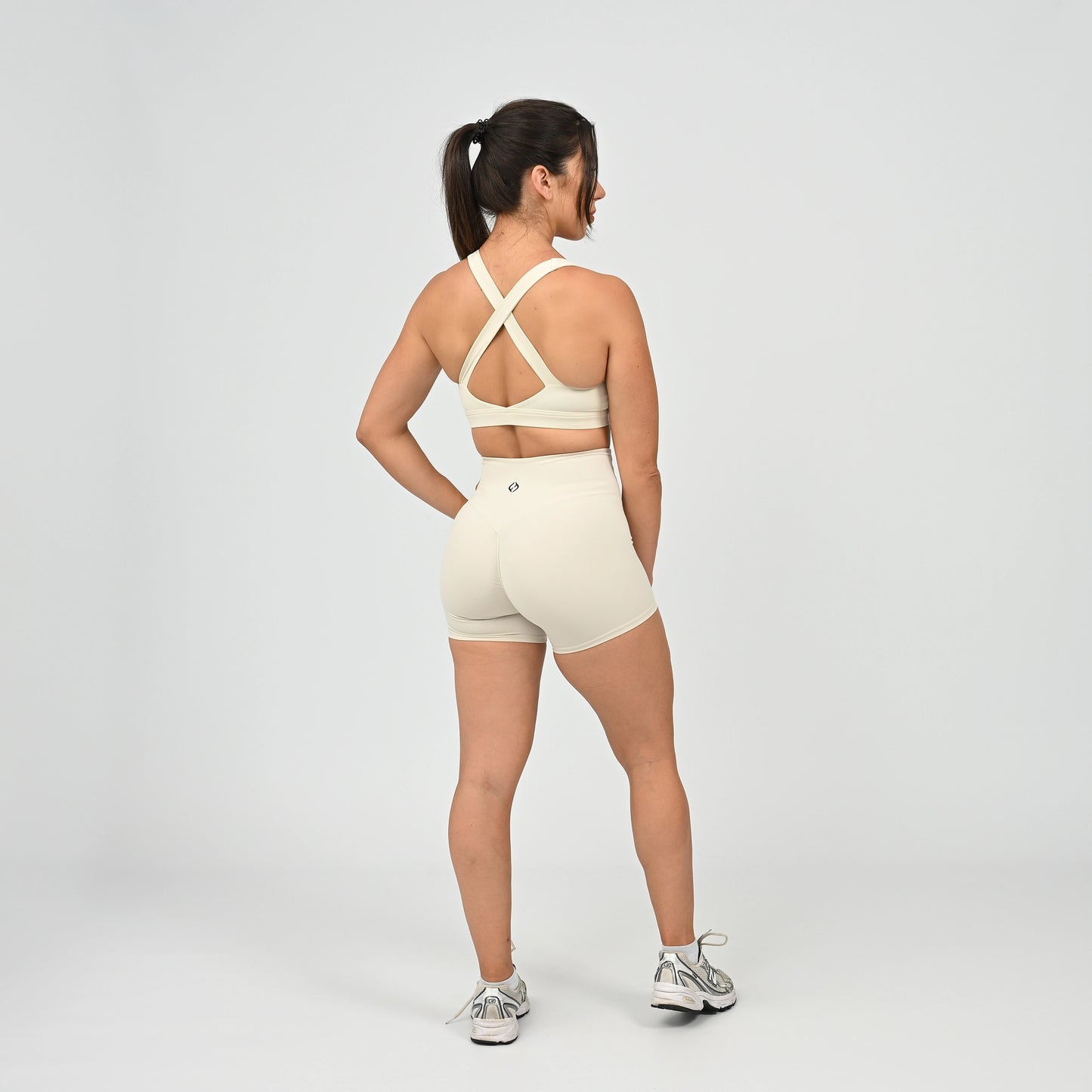 A woman is shown from the back wearing a matching cream-colored activewear set consisting of a sports bra with crisscross straps and high-waisted shorts, paired with gray and white sneakers, against a light gray studio background. From gymwear brand Hybryd.