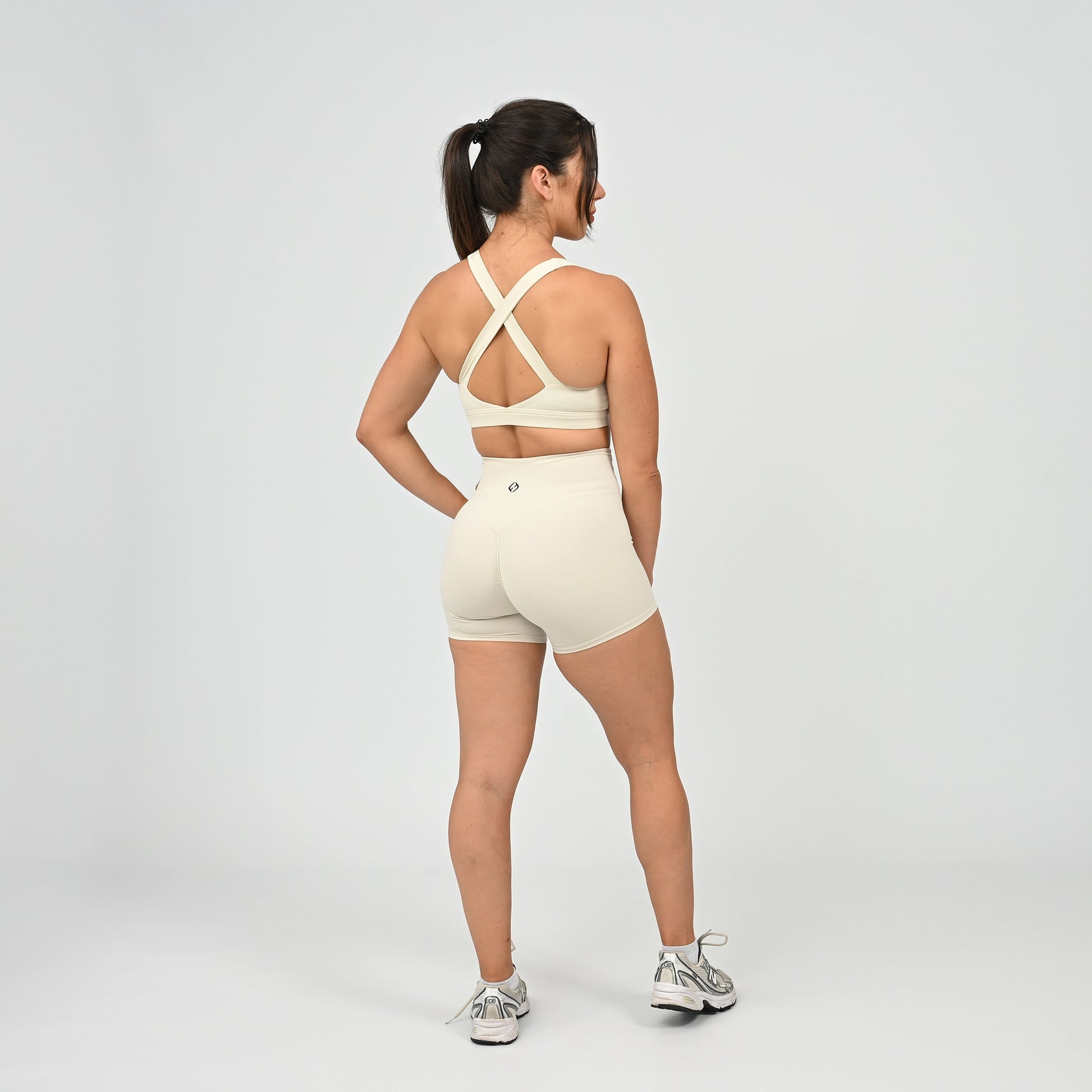 A woman is shown from the back wearing a matching cream-colored activewear set consisting of a sports bra with crisscross straps and high-waisted shorts, paired with gray and white sneakers, against a light gray studio background. From gymwear brand Hybryd.