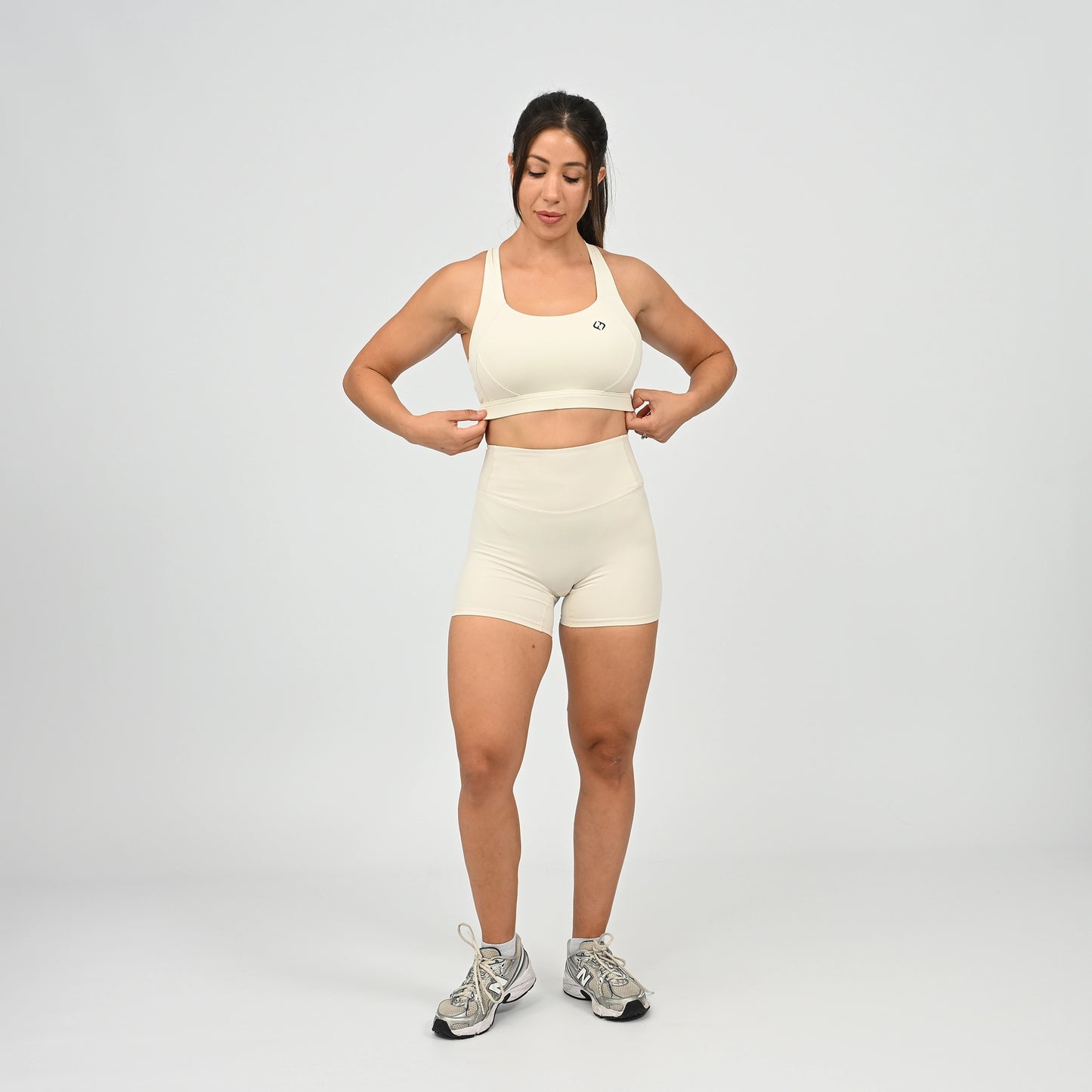 A woman modeling a matching off-white athletic set consisting of a sports bra and high-waisted shorts, paired with gray and white sneakers, against a light gray studio background. From activewear brand Hybryd