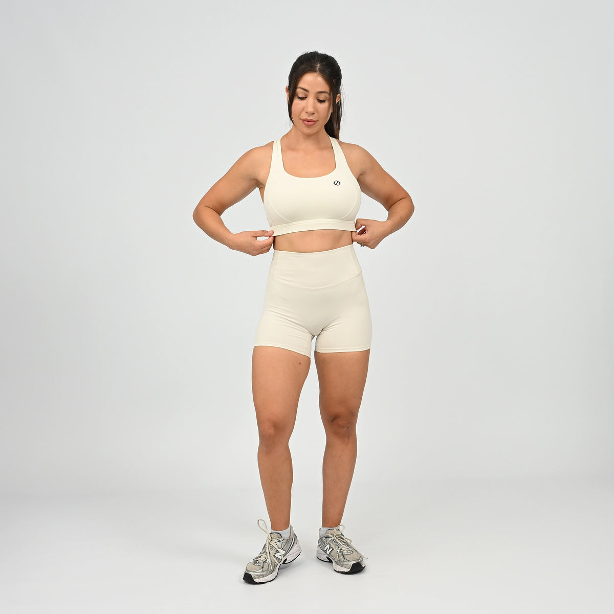 A woman modeling a matching off-white athletic set consisting of a sports bra and high-waisted shorts, paired with gray and white sneakers, against a light gray studio background. From activewear brand Hybryd