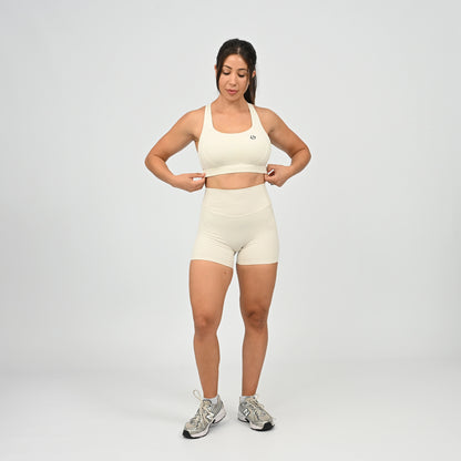 A woman modeling a matching off-white athletic set consisting of a sports bra and high-waisted shorts, paired with gray and white sneakers, against a light gray studio background. From activewear brand Hybryd