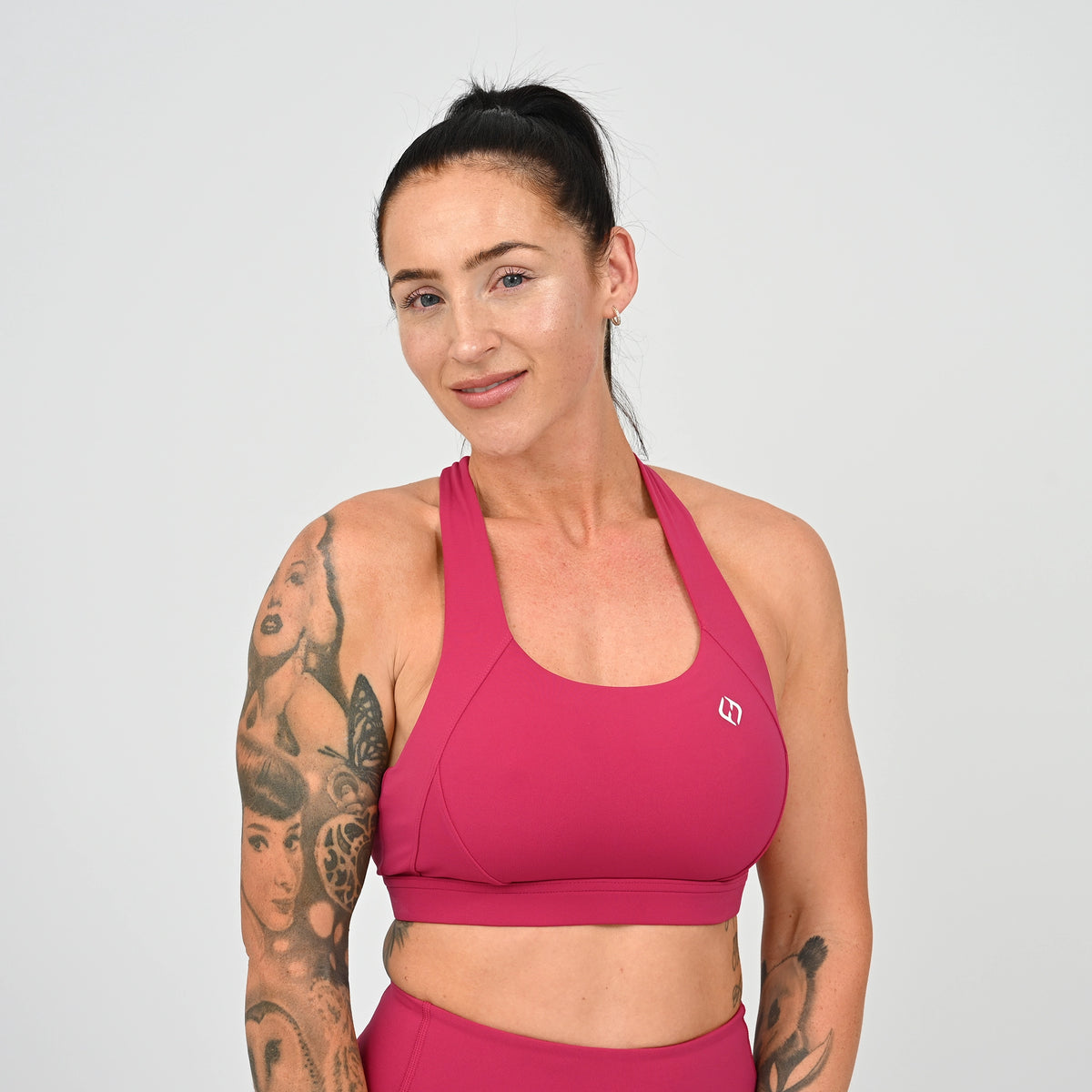 A woman with tattoos modeling a bright raspberry athletic sports bra and matching high-waisted shorts against a light gray studio background. From Hybryd a sports wear brand