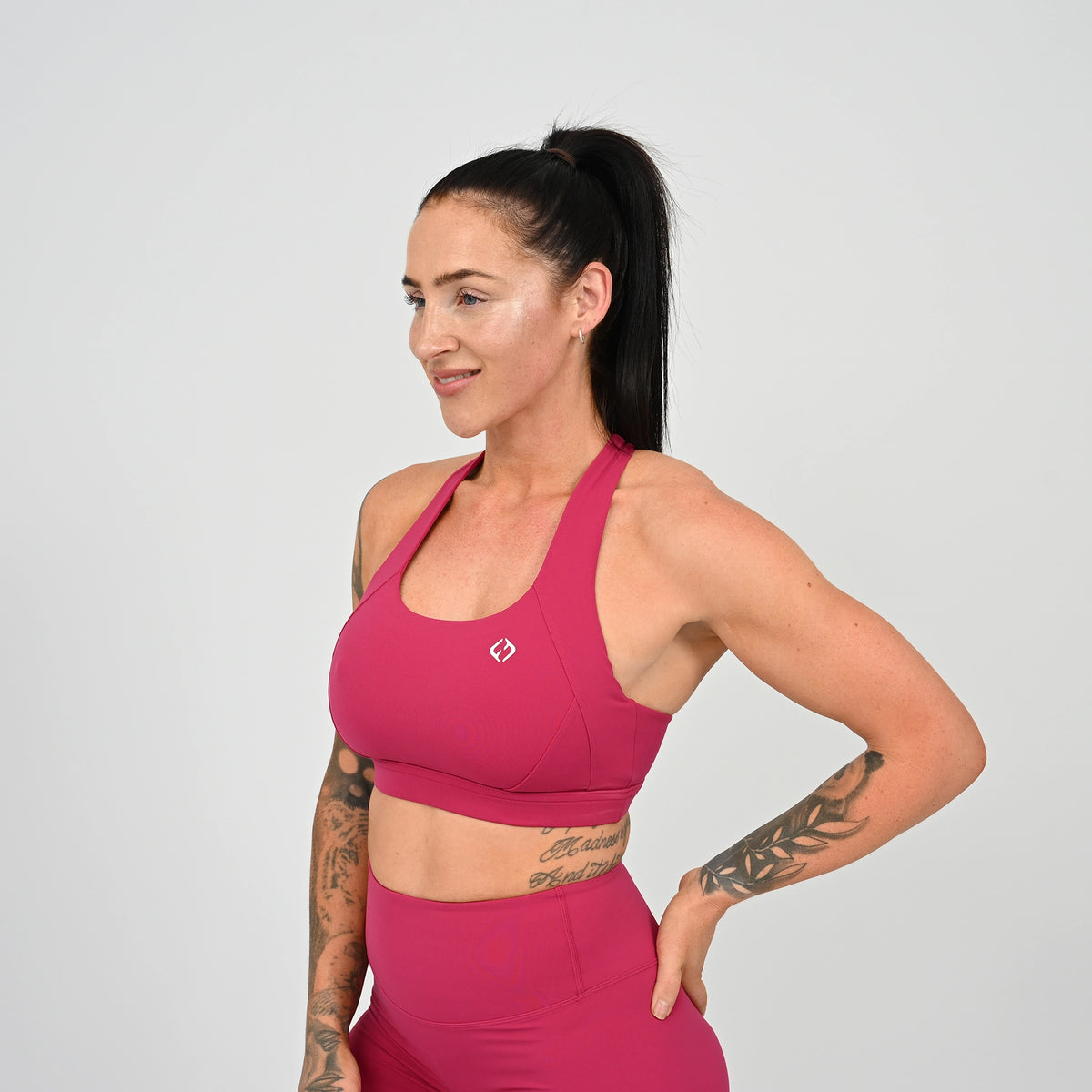 A woman with a ponytail is wearing a raspberry Hybryd sports bra and matching high-waisted shorts against a light gray studio background.