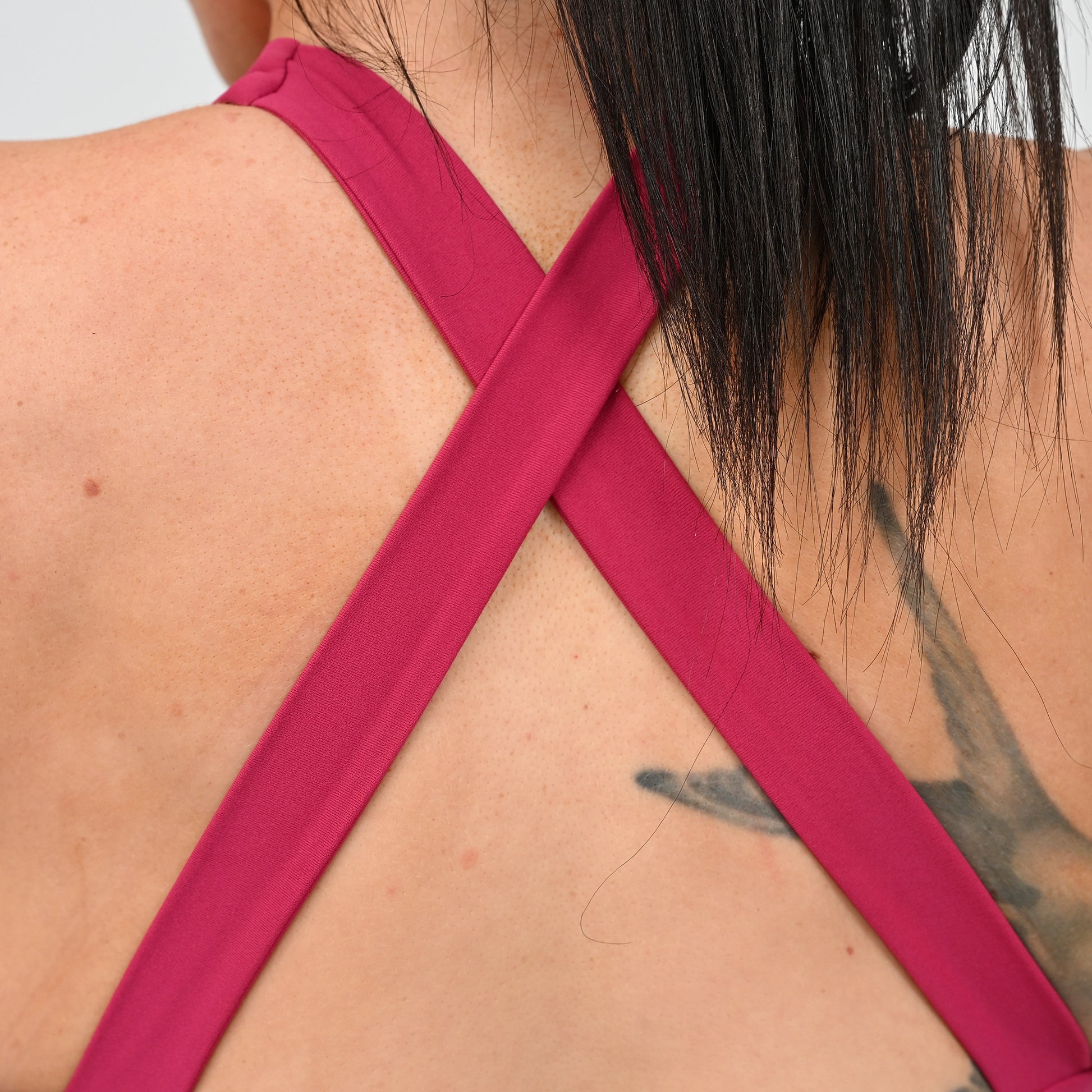 Close-up back view of a woman wearing a bright pink 2 strap bra with wide straps crossing in an X shape. From gymwear brand Hybryd.