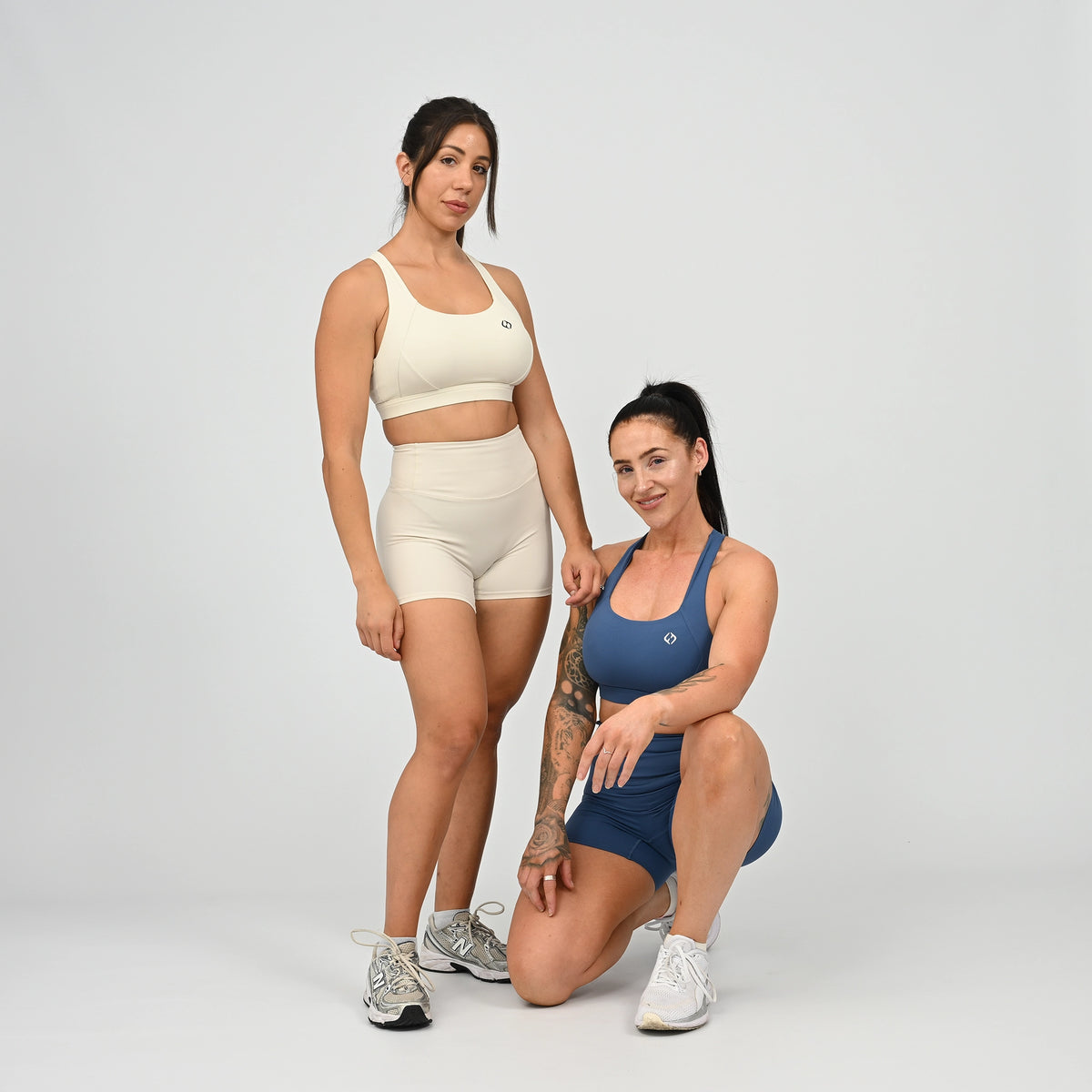 Two women modeling matching Hybryd sets in a studio setting. One woman stands wearing a cream sports bra and shorts set, and the other crouches wearing a blue sports bra and shorts set against a light gray background. From activewear brand Hybryd.
