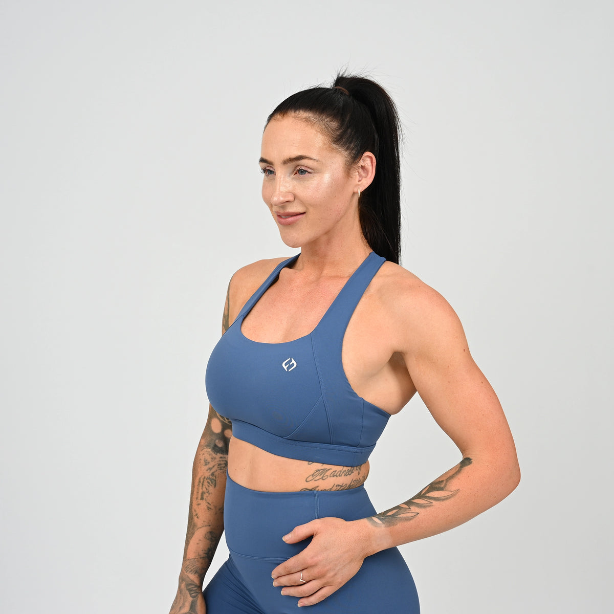 A woman with a ponytail models a matching slate blue athletic set, featuring a sports bra and high-waisted shorts, against a solid light gray background. ll items from activewear brand Hybryd.