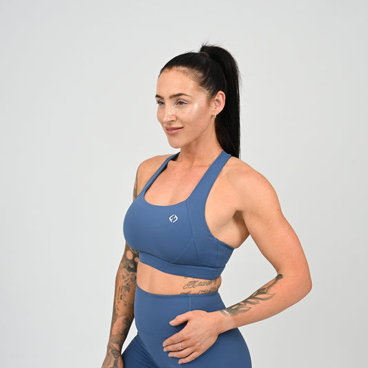 A woman with a ponytail models a matching slate blue athletic set, featuring a sports bra and high-waisted shorts, against a solid light gray background. ll items from activewear brand Hybryd.