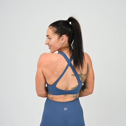 A CrossFit athlete with a high ponytail is shown from the back wearing a blue athletic sports bra and matching shorts against a light gray studio background. From gymwear brand Hybryd.
