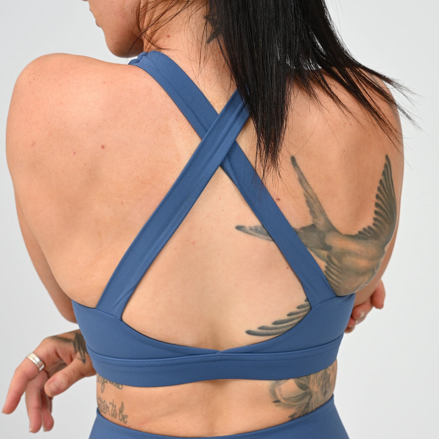 A Hyox athlete shown from the back wearing a blue sports bra with crisscross straps, against a light gray studio background. From gymwear brand Hybryd.