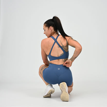 A woman in a blue athletic matching set, consisting of a sports bra and high-waisted shorts, crouching from behind to show the back design of the clothing. She is photographed in a studio setting against a light gray background. From activewear brand Hybryd.