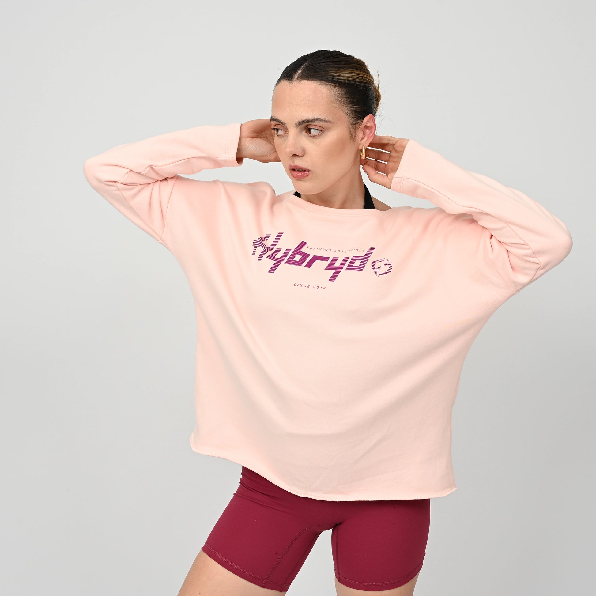 Woman wearing a light pink long-sleeve shirt with 'hybryd' logo on a gray background