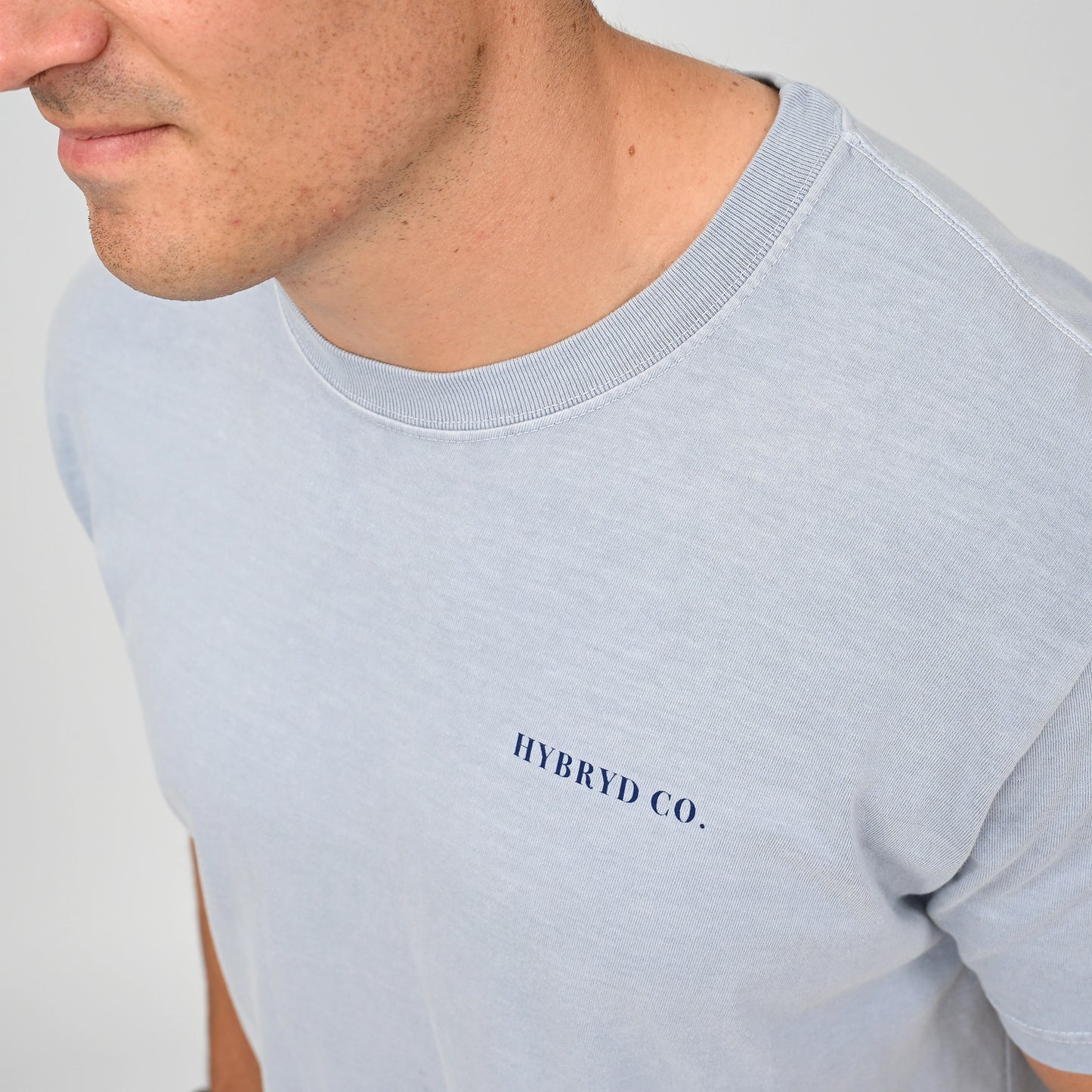Close-up of a man wearing a light blue crewneck t-shirt with 'HYBRYD CO.' printed in navy blue on the chest, against a plain light gray background. Made by gymwear brand Hybryd.