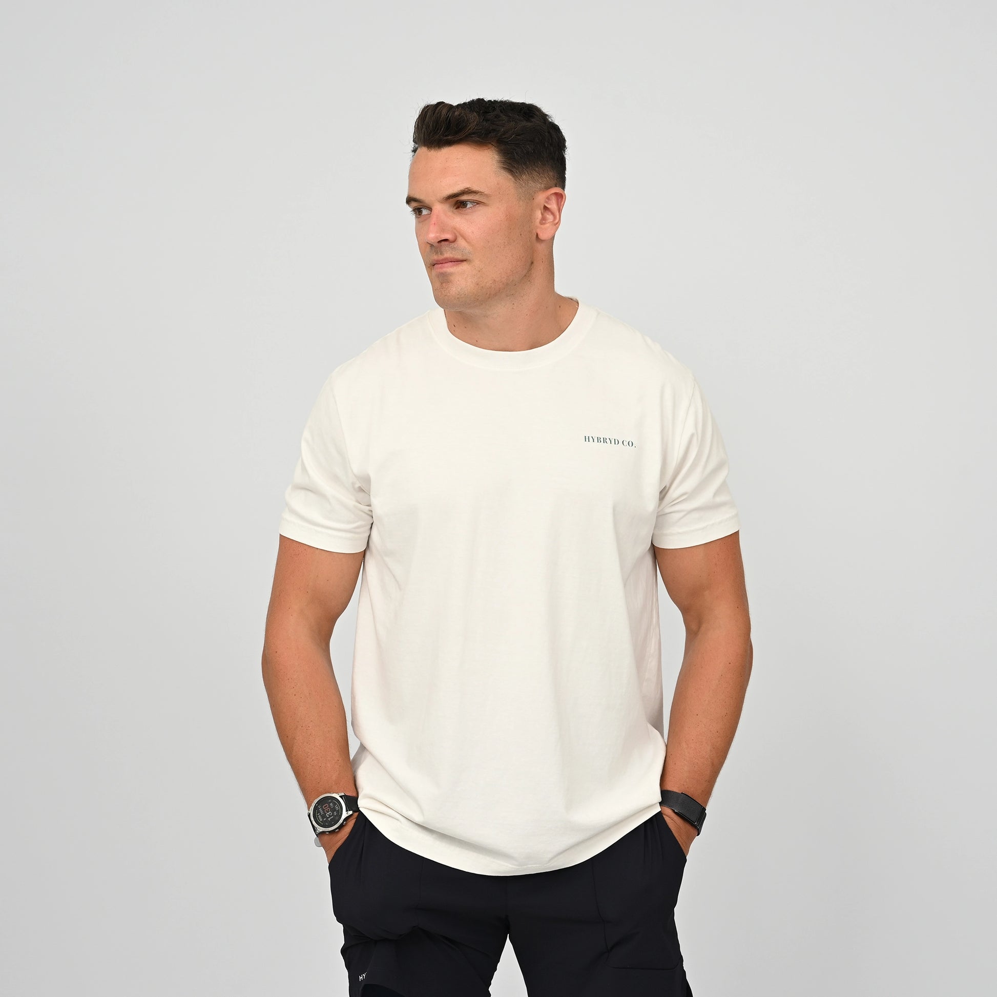 A man wearing a white short-sleeve cotton crewneck t-shirt with a small 'hybryd' logo on the left chest, paired with dark pants, standing against a light gray studio background. From activewear brand Hybryd
