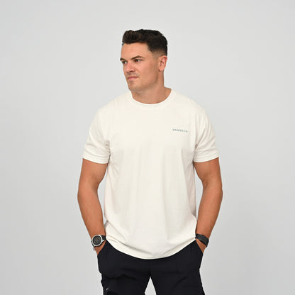 A man wearing a white short-sleeve cotton crewneck t-shirt with a small 'hybryd' logo on the left chest, paired with dark pants, standing against a light gray studio background. From activewear brand Hybryd