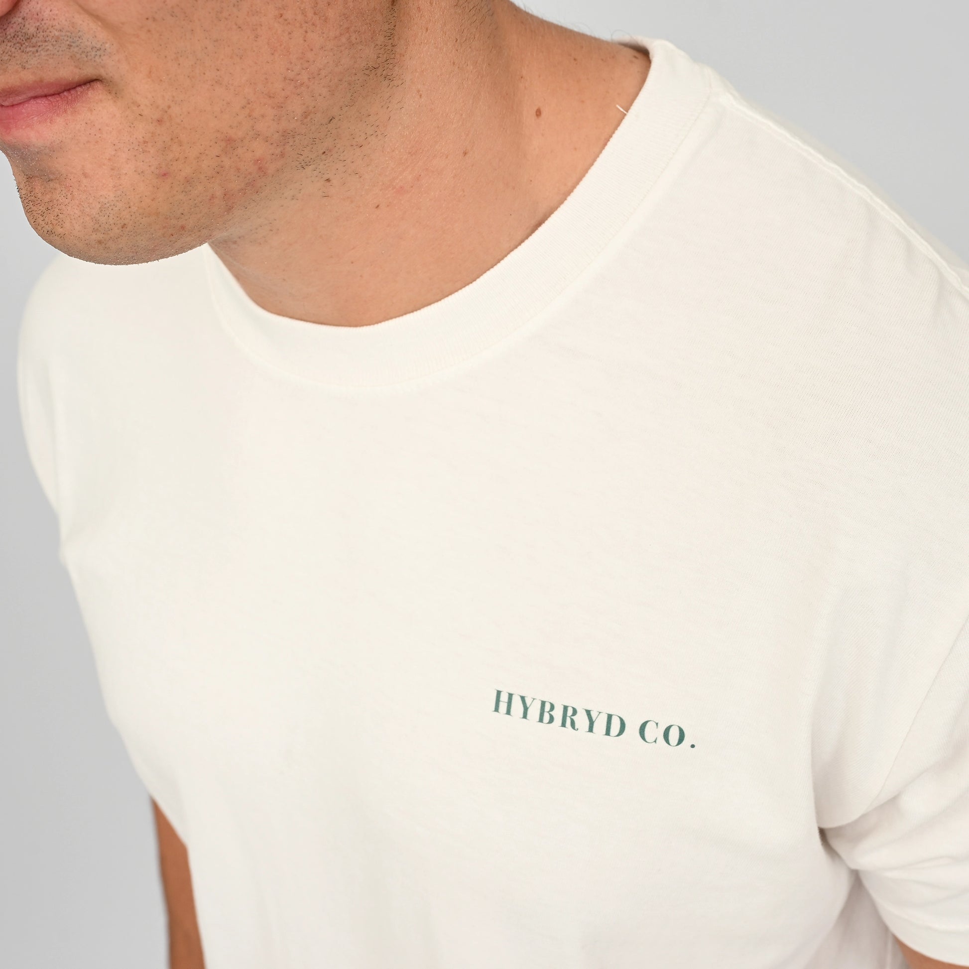 Close-up of a man wearing an off-white crewneck t-shirt with 'HYBRYD CO.' printed in dark green on the chest, set against a neutral gray background. From actiewear brand Hybryd.