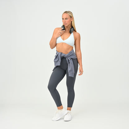 A woman modeling dark gray athletic leggings with a Hybryd jumper tied around her waist, a white Hybryd sports bra, and white sneakers, standing against a light gray studio background.